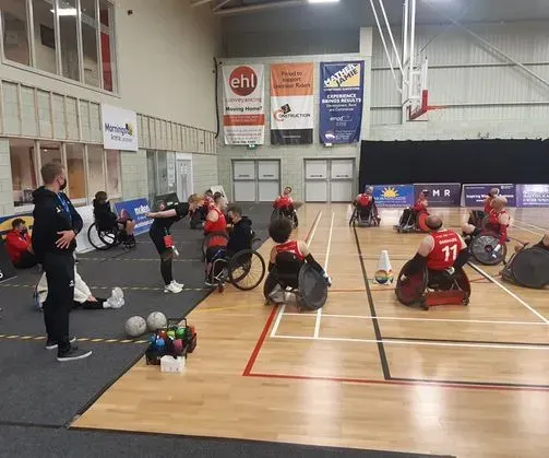 Kørestolsrugbytræning i en sportshal. Spillere i røde trøjer, træner instruerer, bolde på gulvet og bannere på væggen.