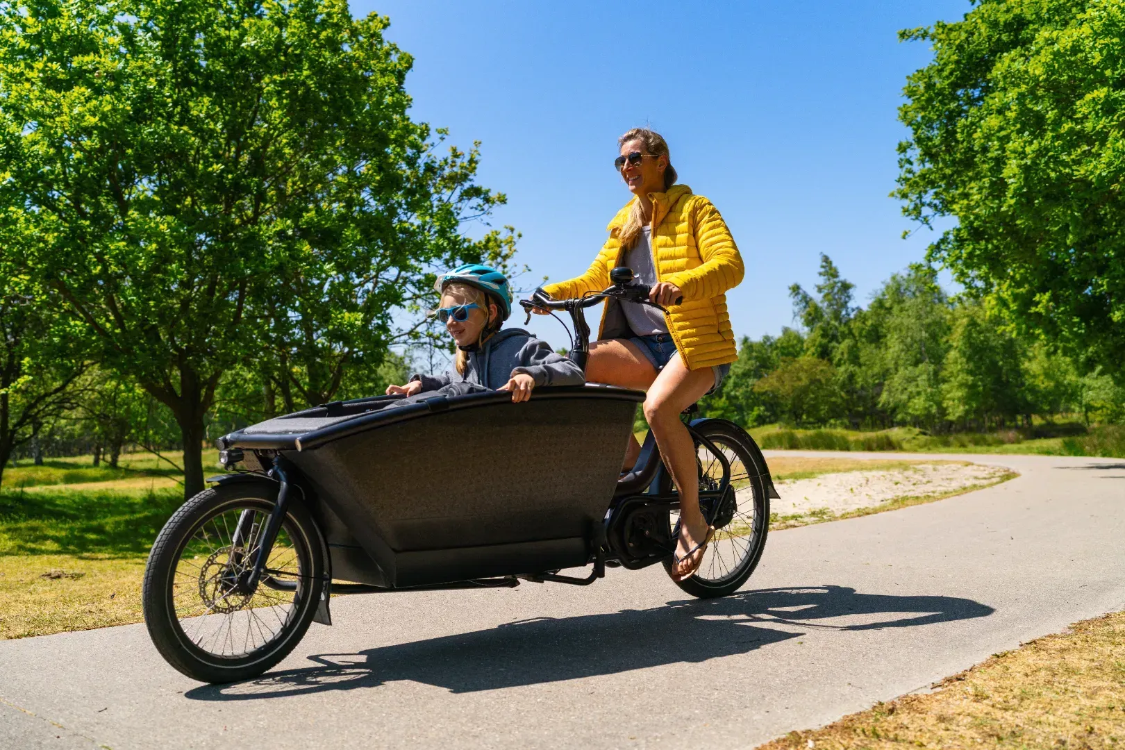Kvinde kører på en lad-elcykel med et barn foran på en asfalteret sti, solskinsdag.
