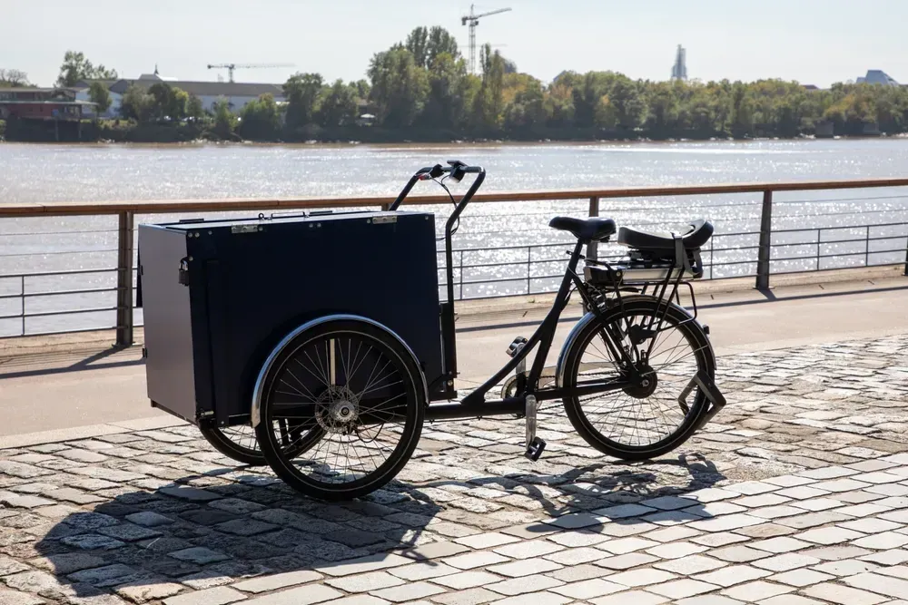 Ladcykel med stor blå kasse foran, parkeret på brostenssti ved vandet.