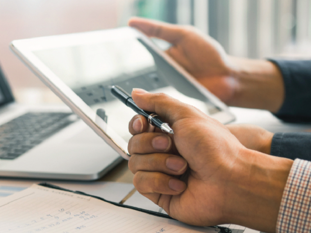 Hands holding a pen and tablet beside a laptop on a desk with charts.