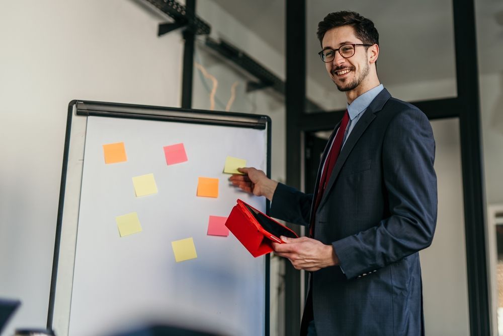 A professional in a suit smiles while pointing to a whiteboard covered with colorful sticky notes in an office setting.