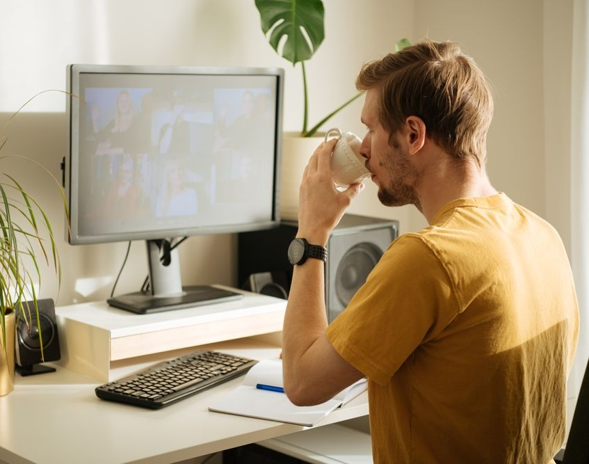 Person at a desk drinking from a mug while looking at a computer monitor in a sunlit home office