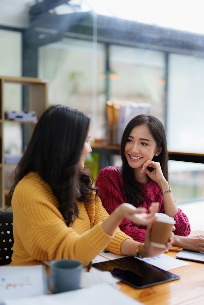 Two women chatting at a breakroom table, smiling with coffee cups and a tablet nearby.