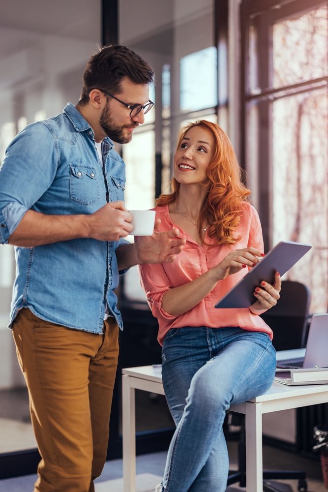 Two coworkers chatting by a desk, one holding a mug and the other a tablet, in a bright office