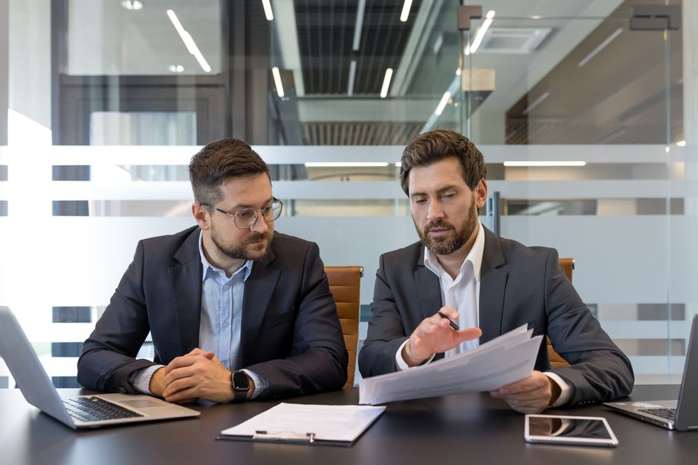 Two businessmen in suits reviewing documents at a conference table in a glass office