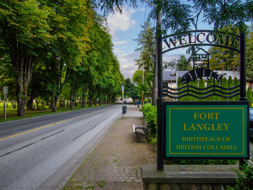 Tree-lined road beside Fort Langley welcome sign and sidewalk under cloudy sky