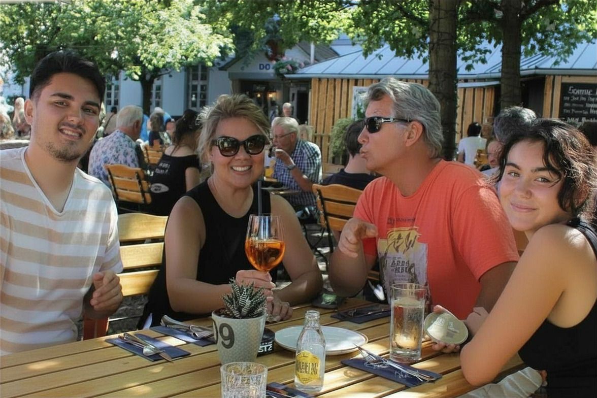 Four friends sit at an outdoor café table, smiling with drinks in sunlight.