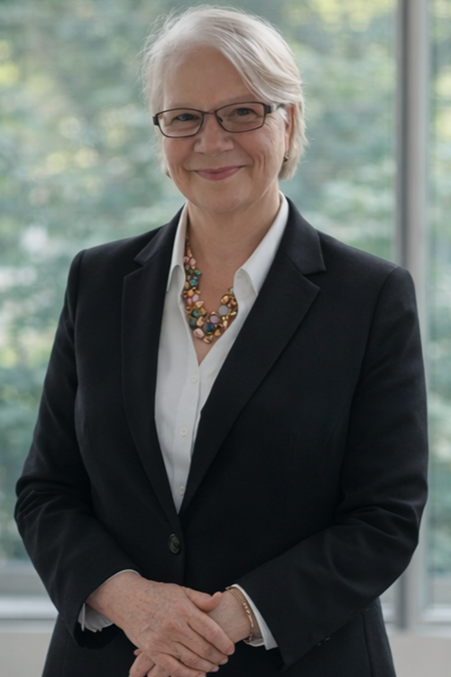 Portrait of a smiling person in a black blazer and white shirt, standing by a window.