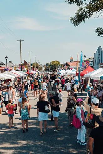 Crowded outdoor festival with white tents, colorful booths, and people walking along a sunlit street.
