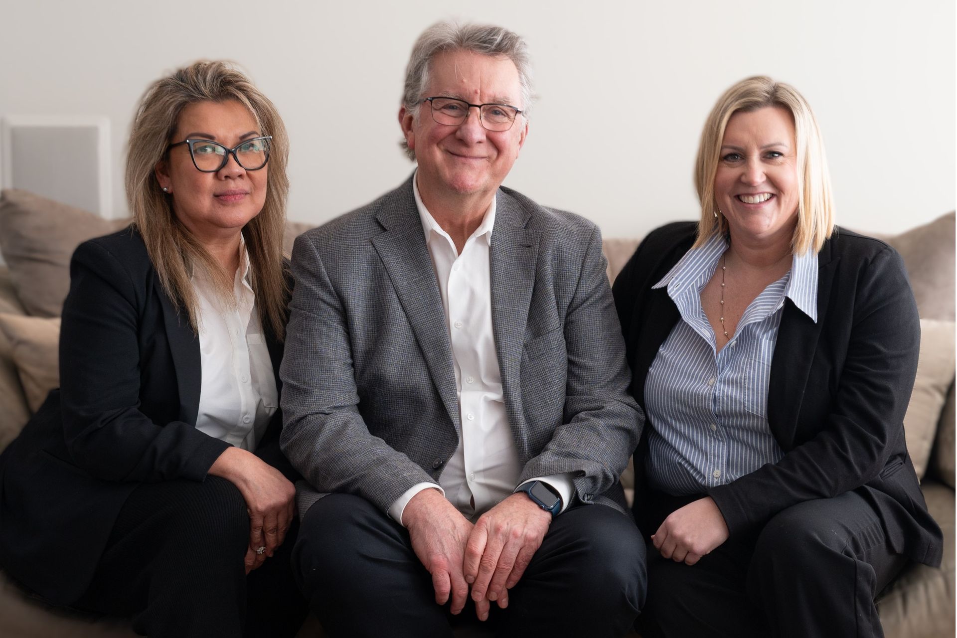 Three smiling professionals seated together on a couch in business attire, posing in a bright office setting.