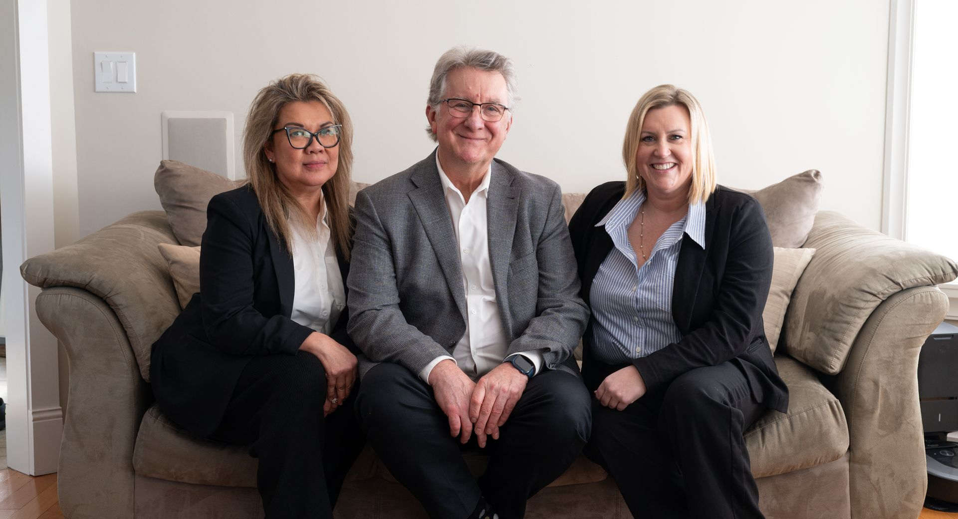 Three people sit smiling on a beige couch in a bright living room.
