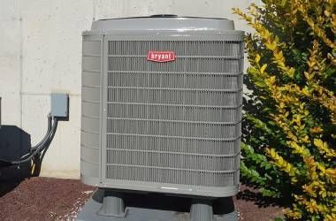 A large gray air conditioner is sitting outside next to a bush.