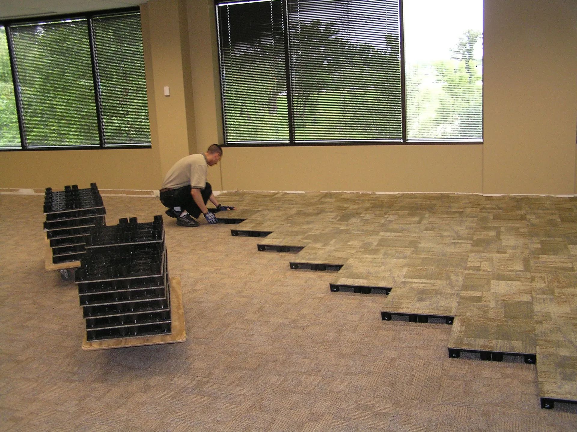 Person crouching beside a row of black crates on a carpeted ramp in a room with large windows.