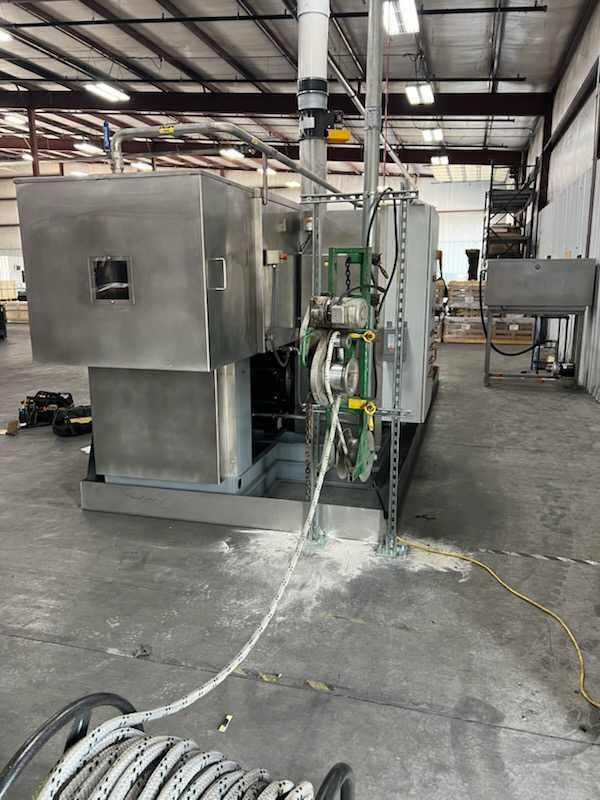 Industrial machine in a warehouse setting, with stainless steel enclosure and connected pipes and wires.