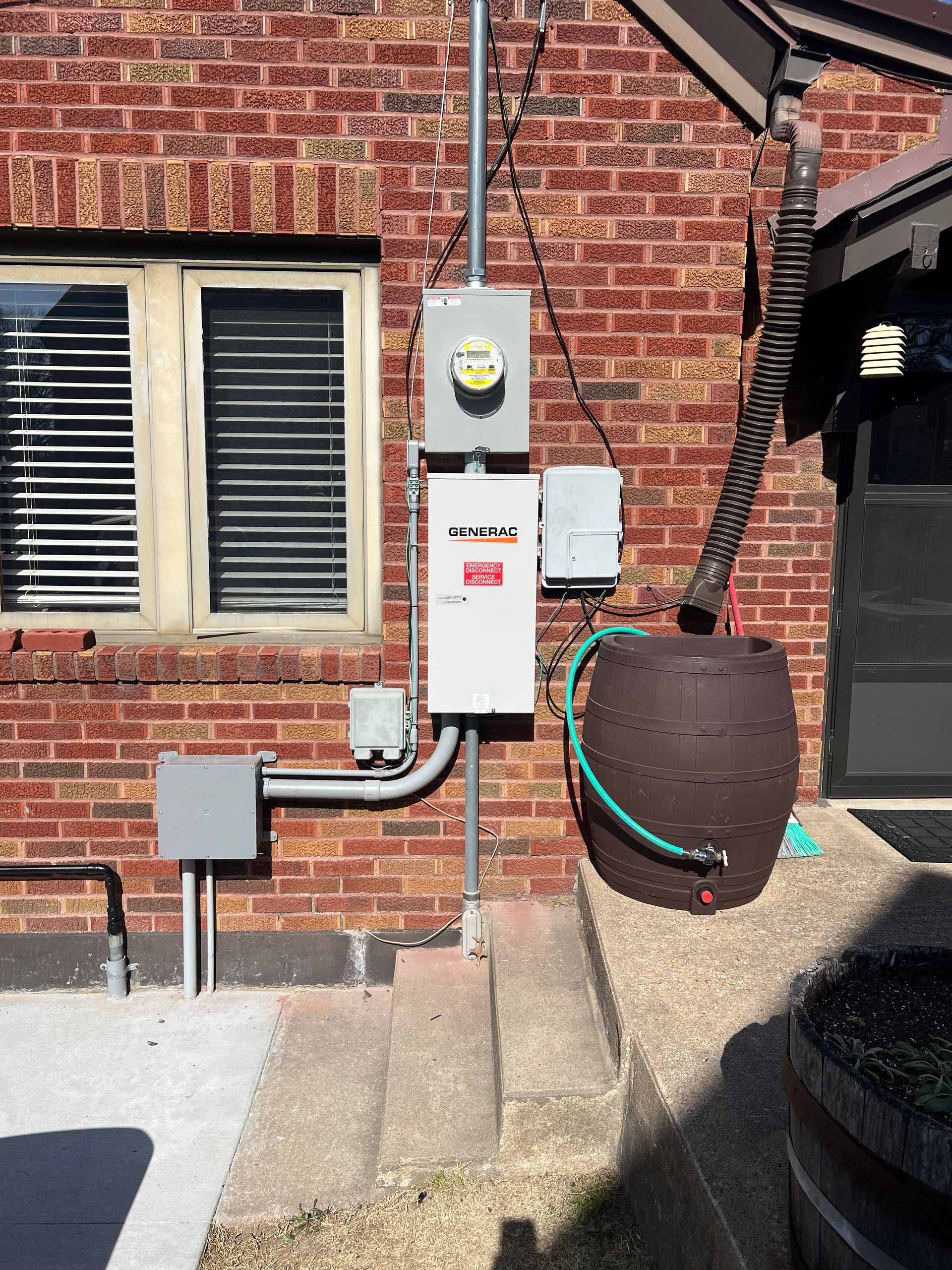Electrical meter and breaker boxes mounted on a brick wall next to a rain barrel and downspout.