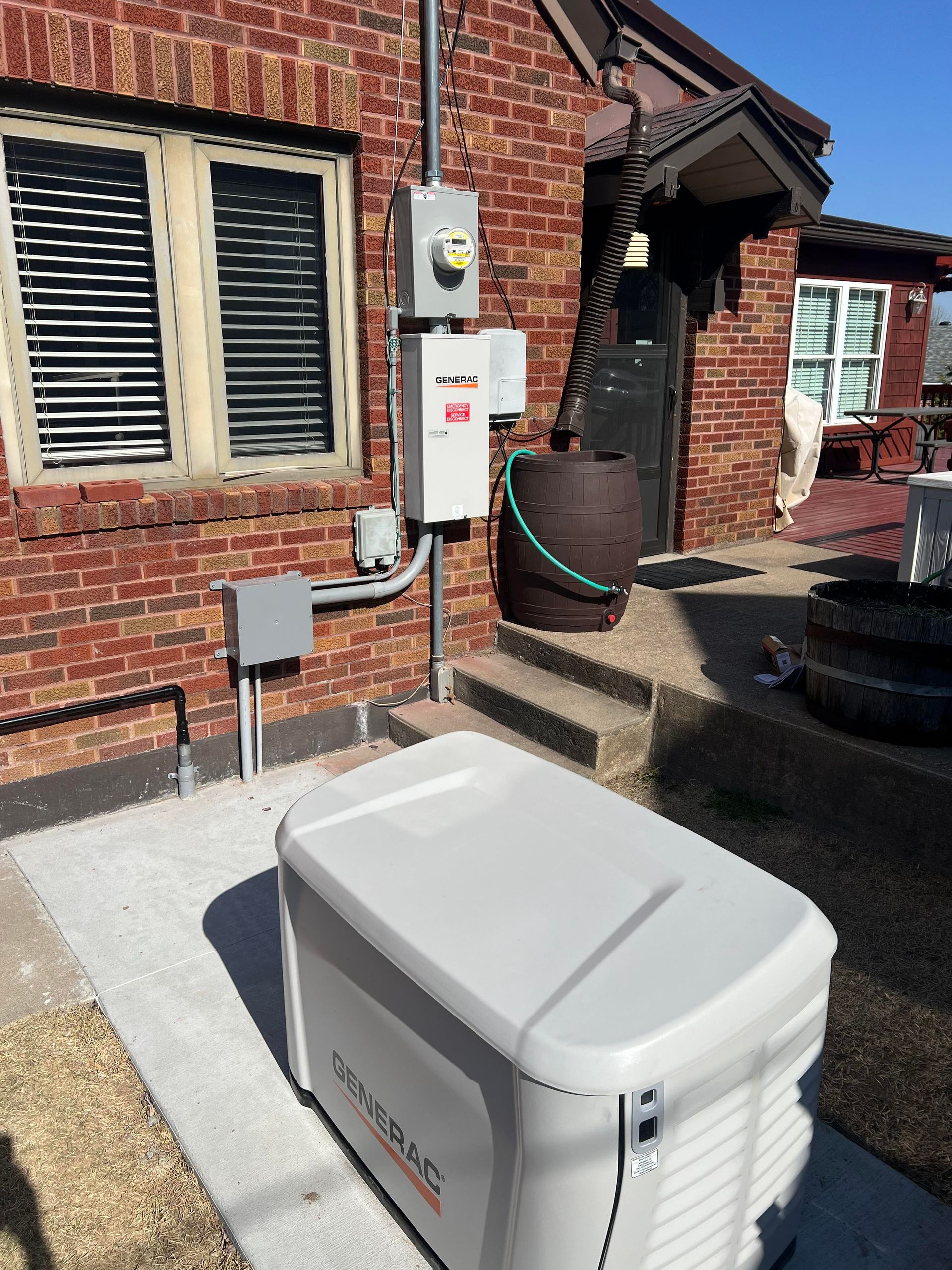 A Generac standby generator installed on a concrete pad next to a brick house with electrical conduit and boxes on the wall.