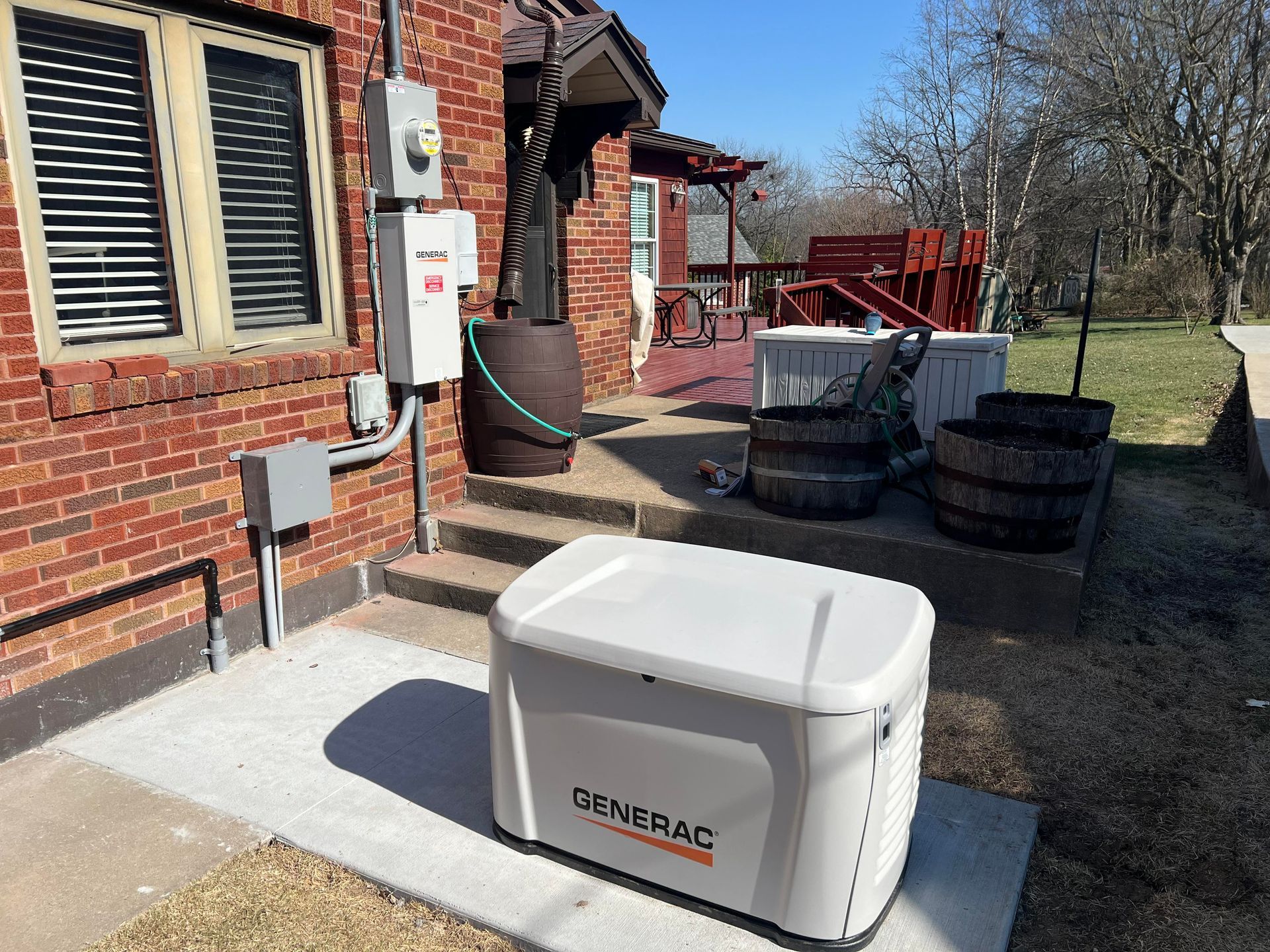 White portable generator on a mat beside a brick house, with a hose and stacked tires in the yard.