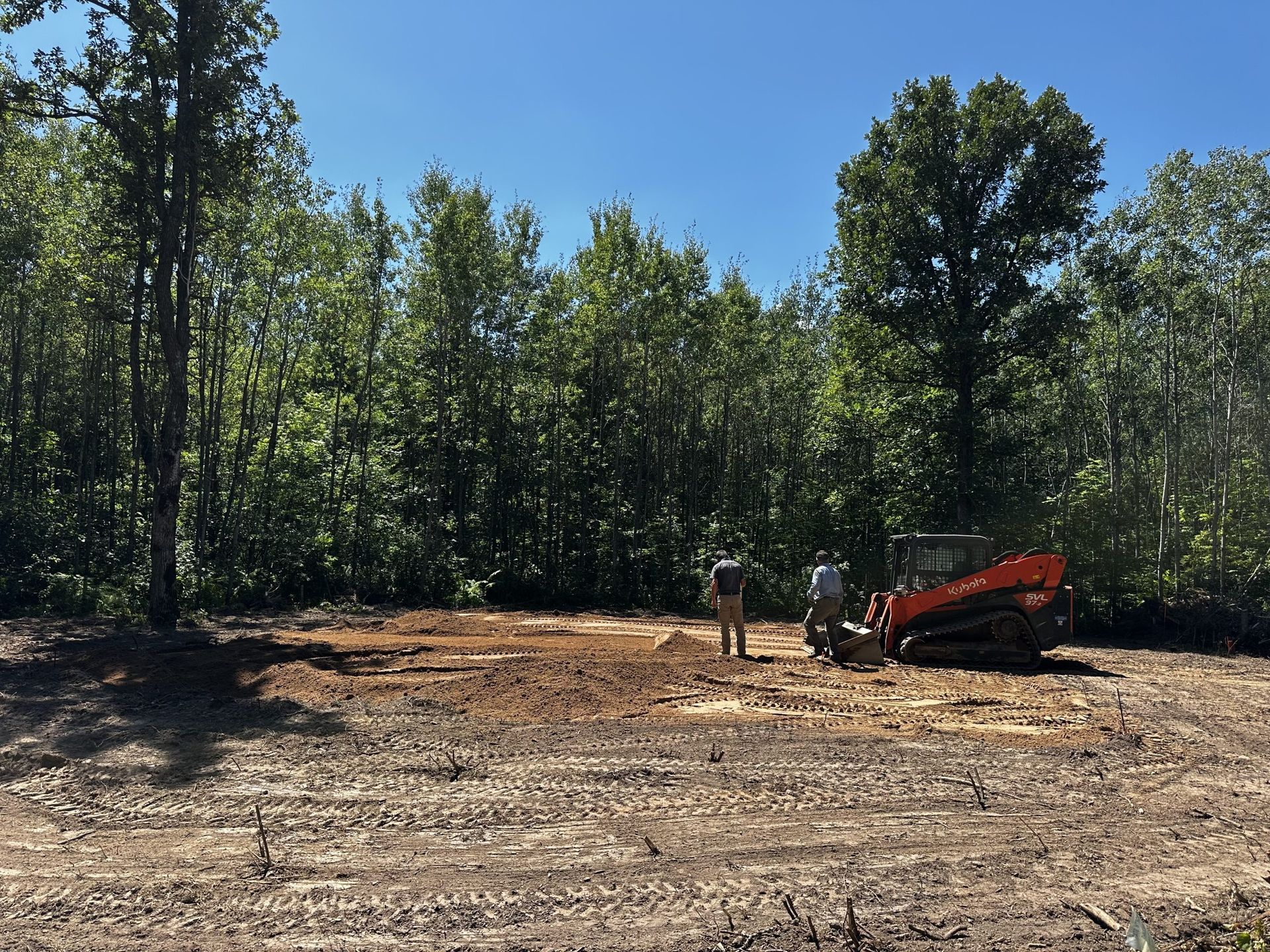 Excavator working on a cleared dirt lot beside a tree line under a blue sky