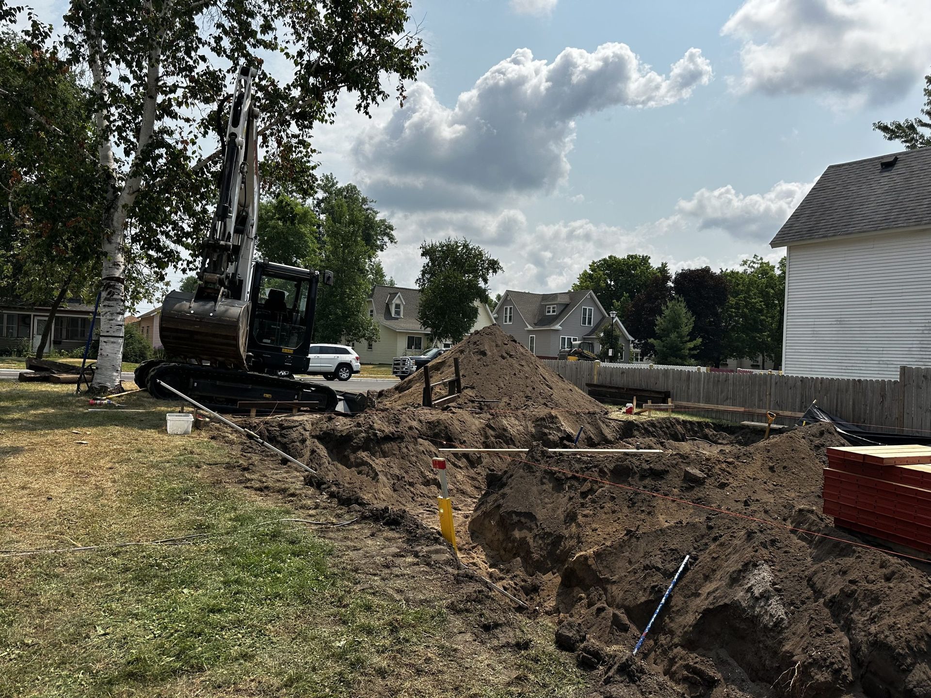 Backyard construction trench with excavator, dirt pile, and a house in the background
