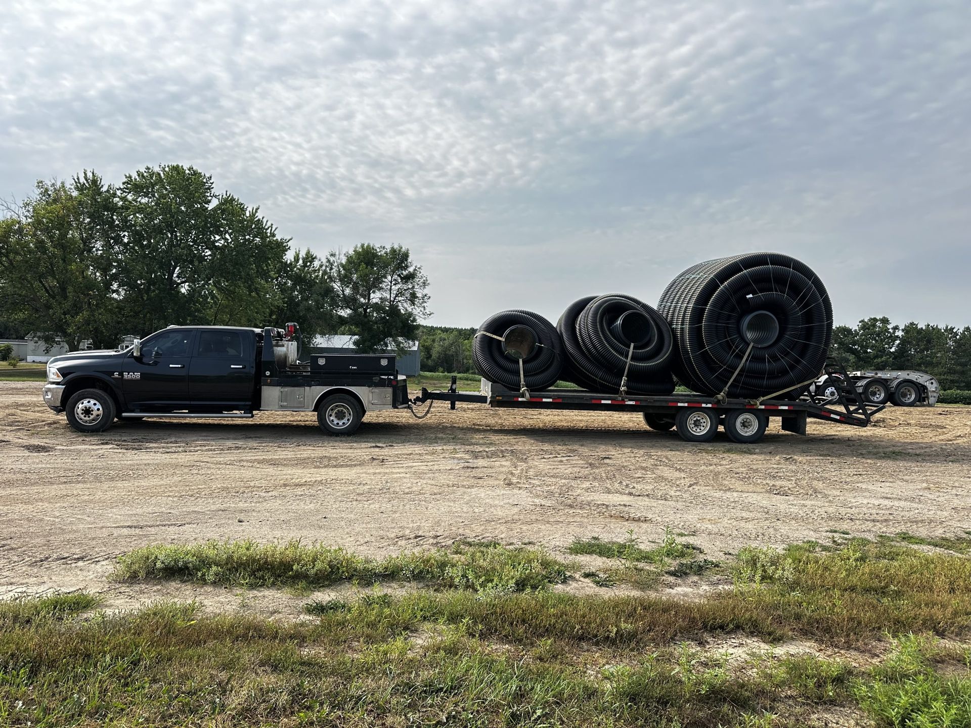 Black pickup truck towing a trailer loaded with large tires on a gravel lot under cloudy skies