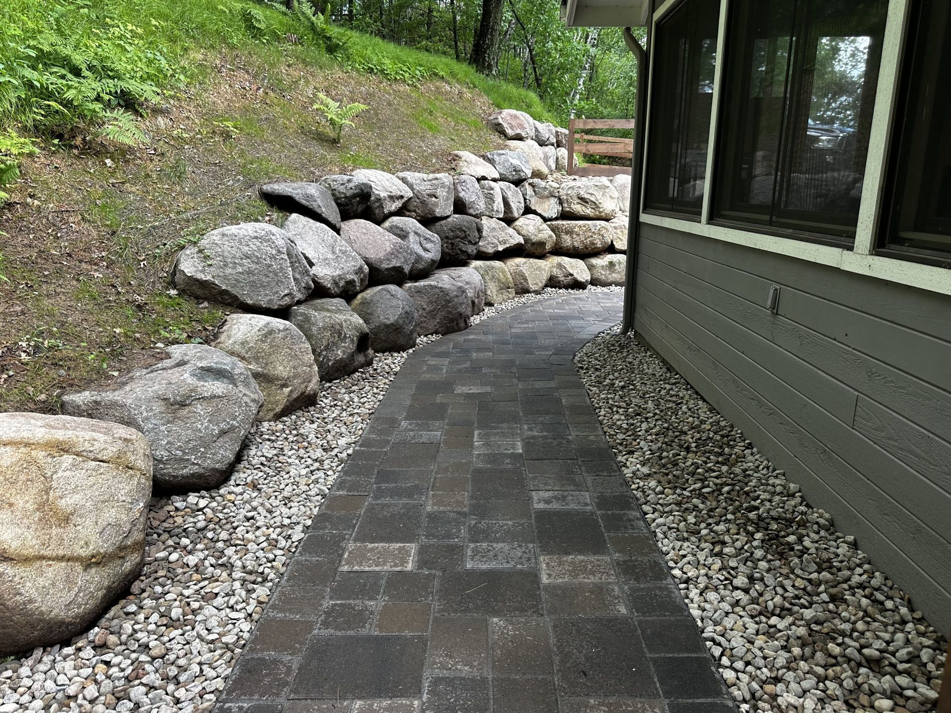 Curved stone walkway beside a house, bordered by gravel and a stacked rock retaining wall.