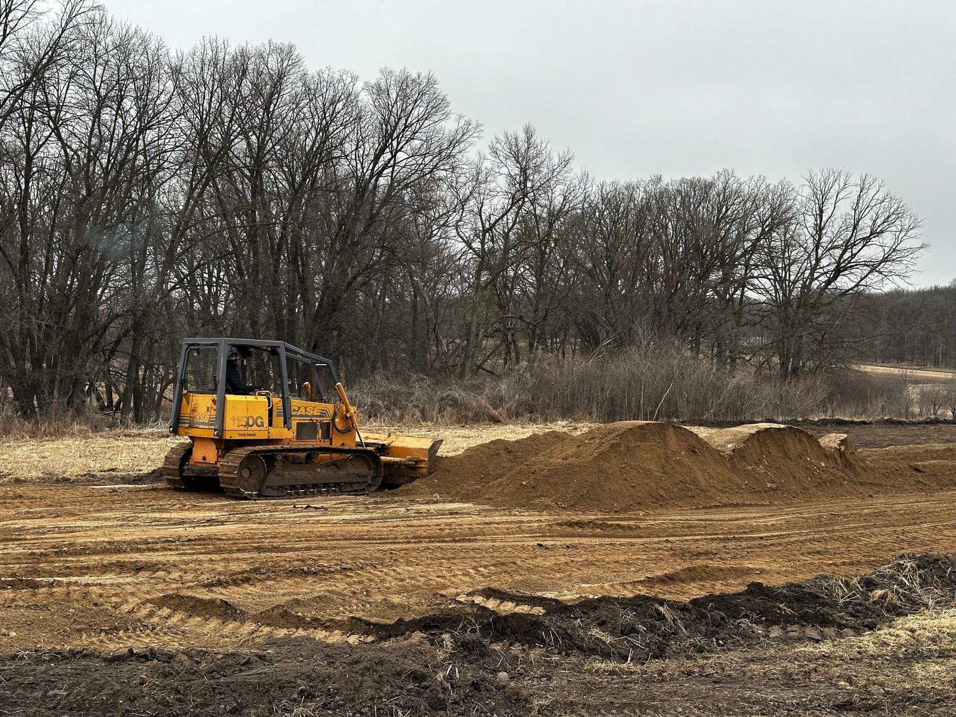 Yellow bulldozer grading a dirt lot with mounds of soil and leafless trees in the background