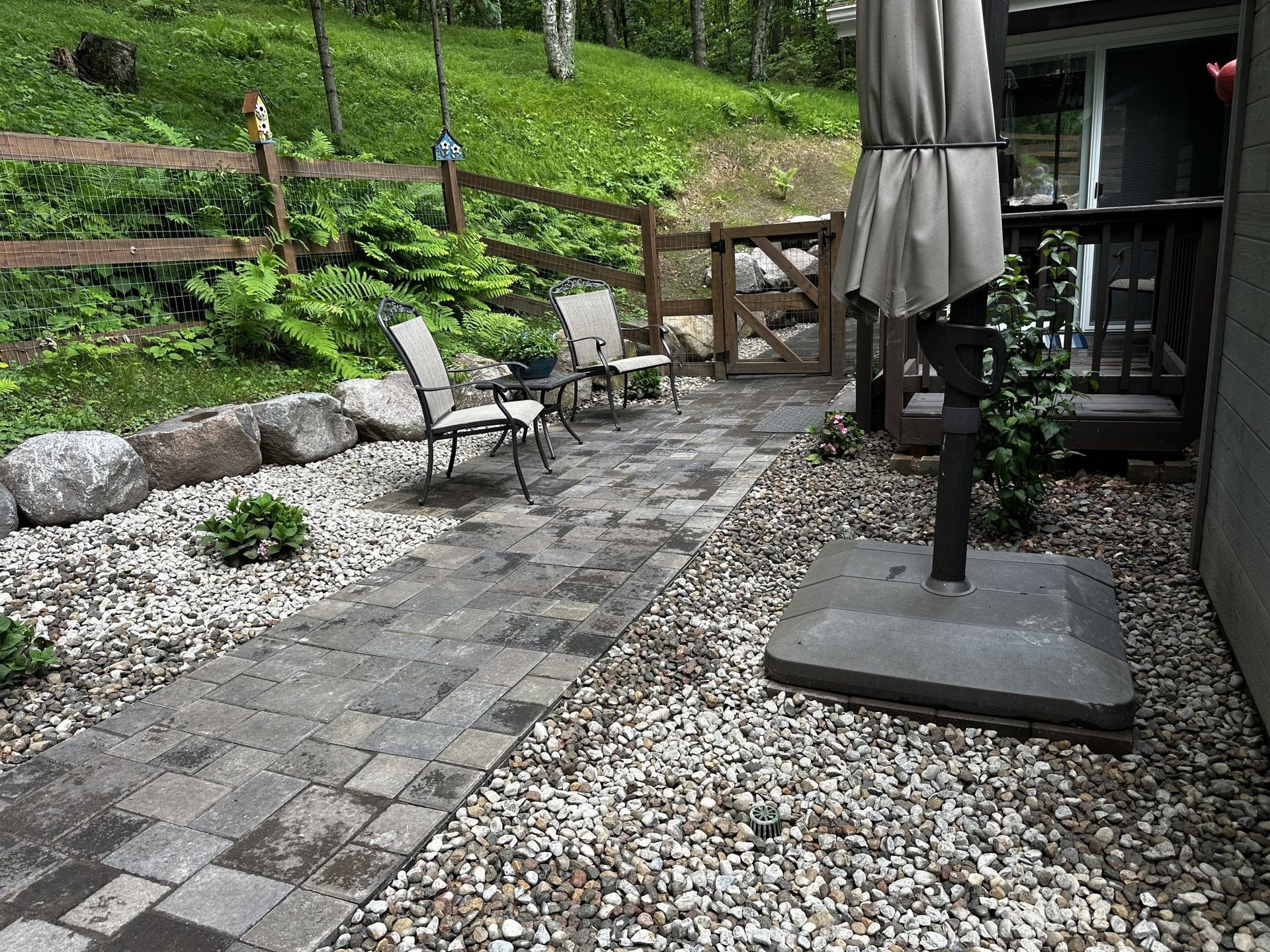 Patio with stone pavers, white chairs, gravel borders, and a closed beige umbrella beside a house