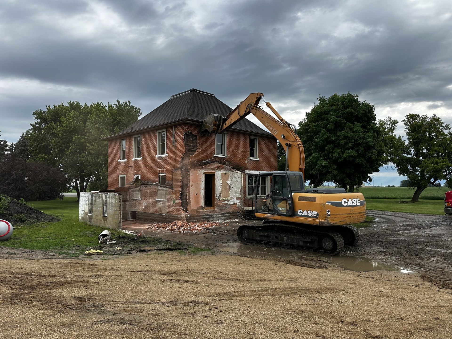 Excavator demolishing a two-story brick house under a cloudy sky, with debris in the yard
