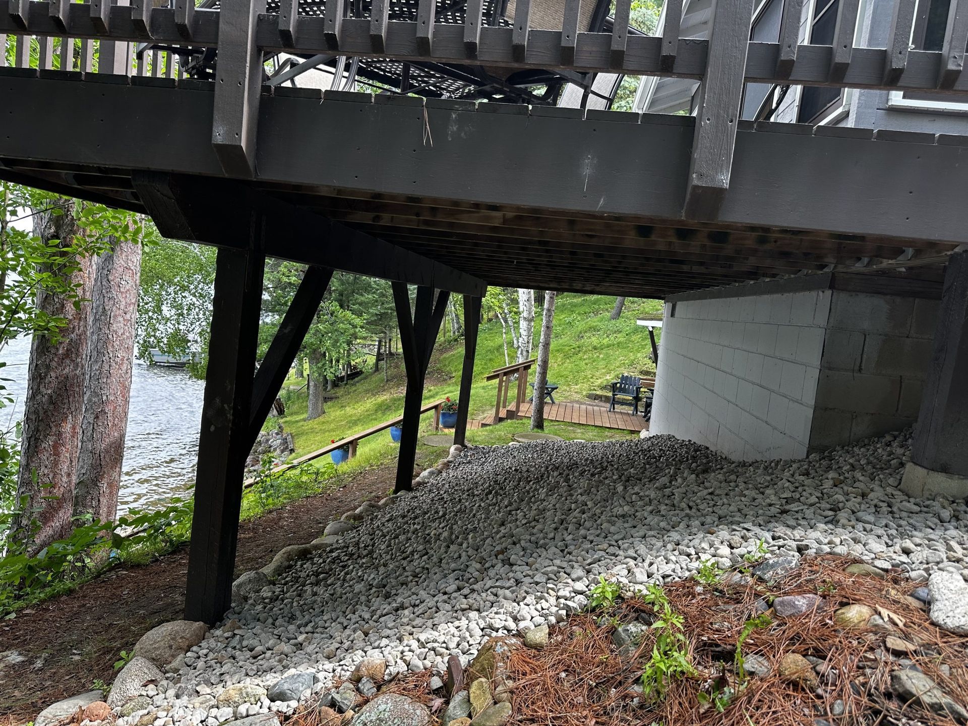 Under a wooden deck beside a lake, with gravel slope, trees, and a black support post.