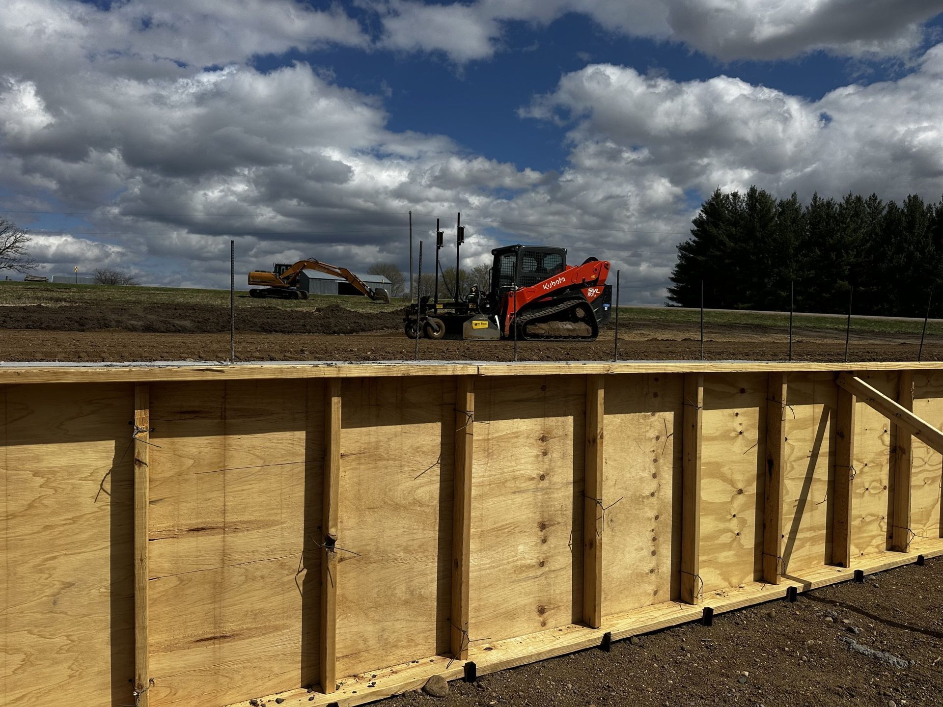 Wooden retaining wall in foreground, with construction equipment and cloudy sky on a worksite behind it.