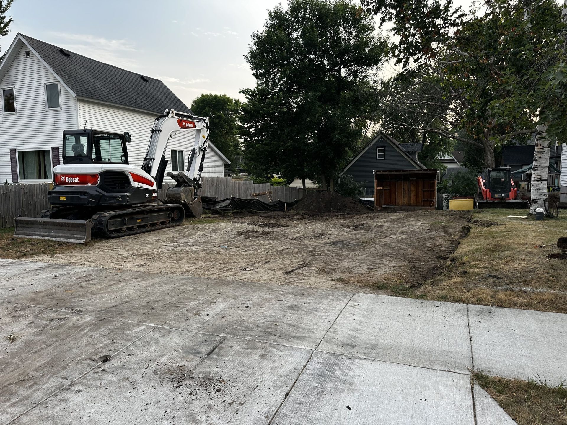 Construction equipment and excavator working on a dirt driveway beside a white house and garage.