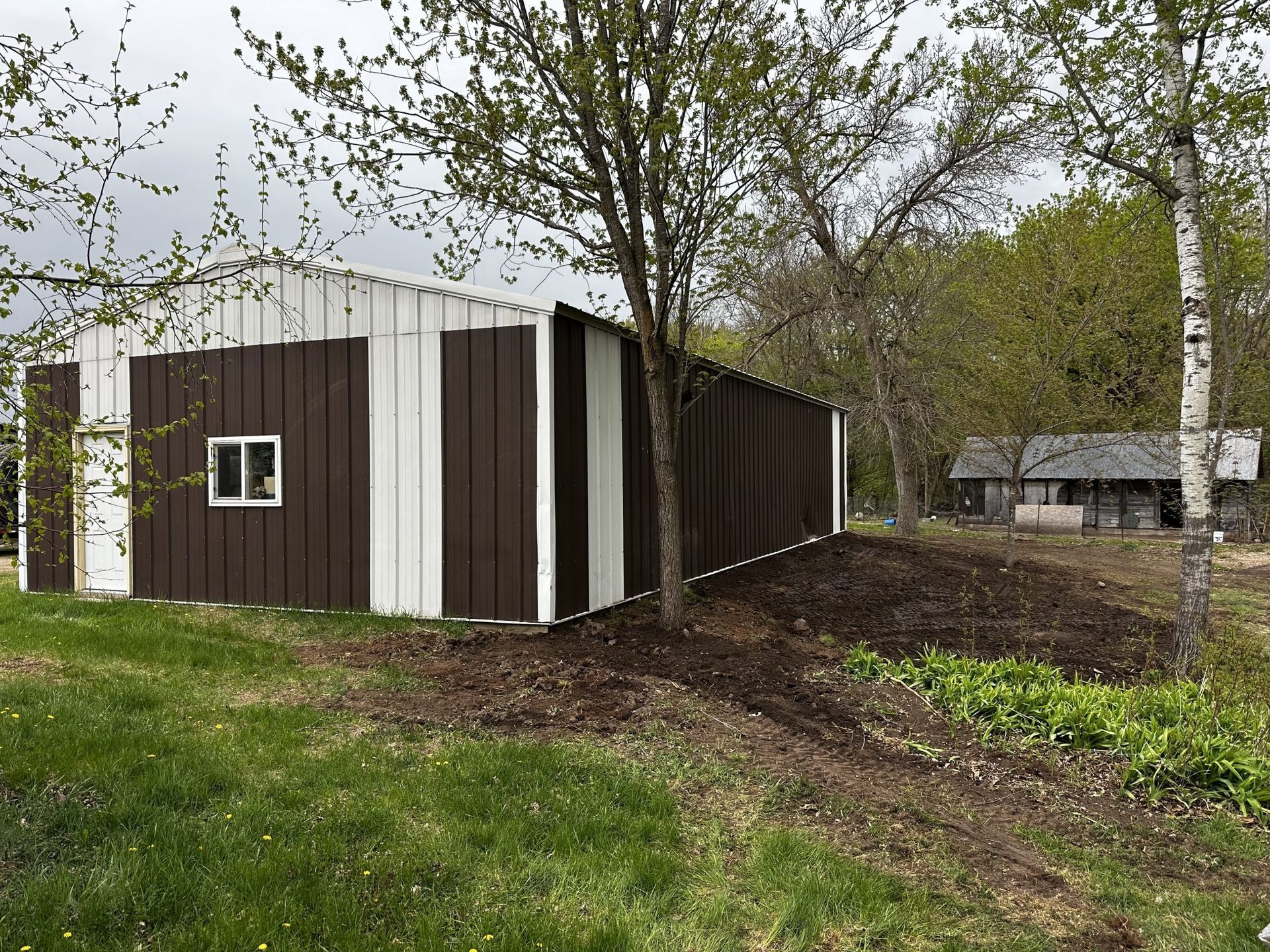 Small brown-and-white shed in a grassy yard with trees and a dirt path nearby