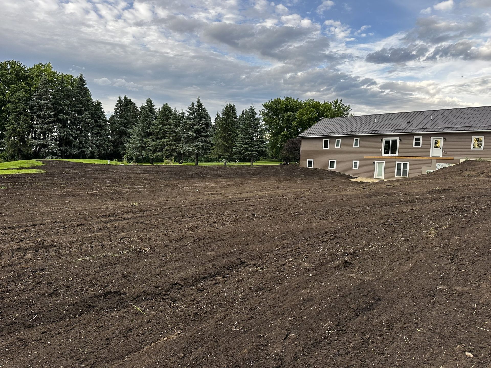 Freshly graded dirt lot beside a beige house, with evergreen trees and a cloudy sky