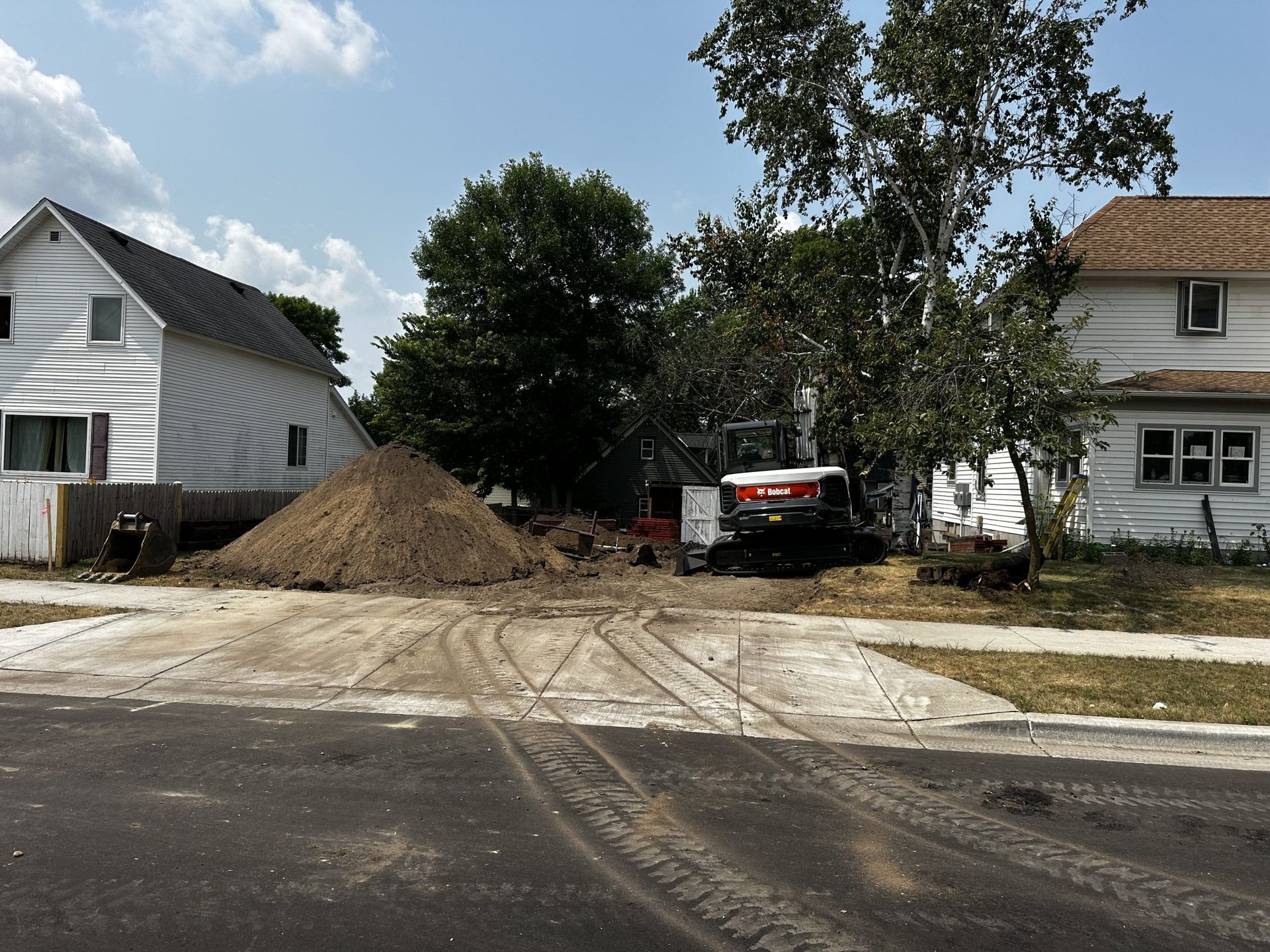 Construction materials and a skid-steer piled in a residential driveway between two white houses