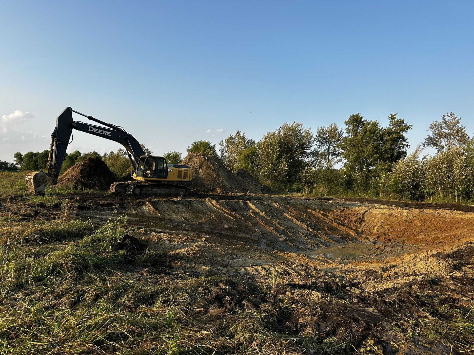 Excavator working on a dirt lot under a clear blue sky, with trees in the background.