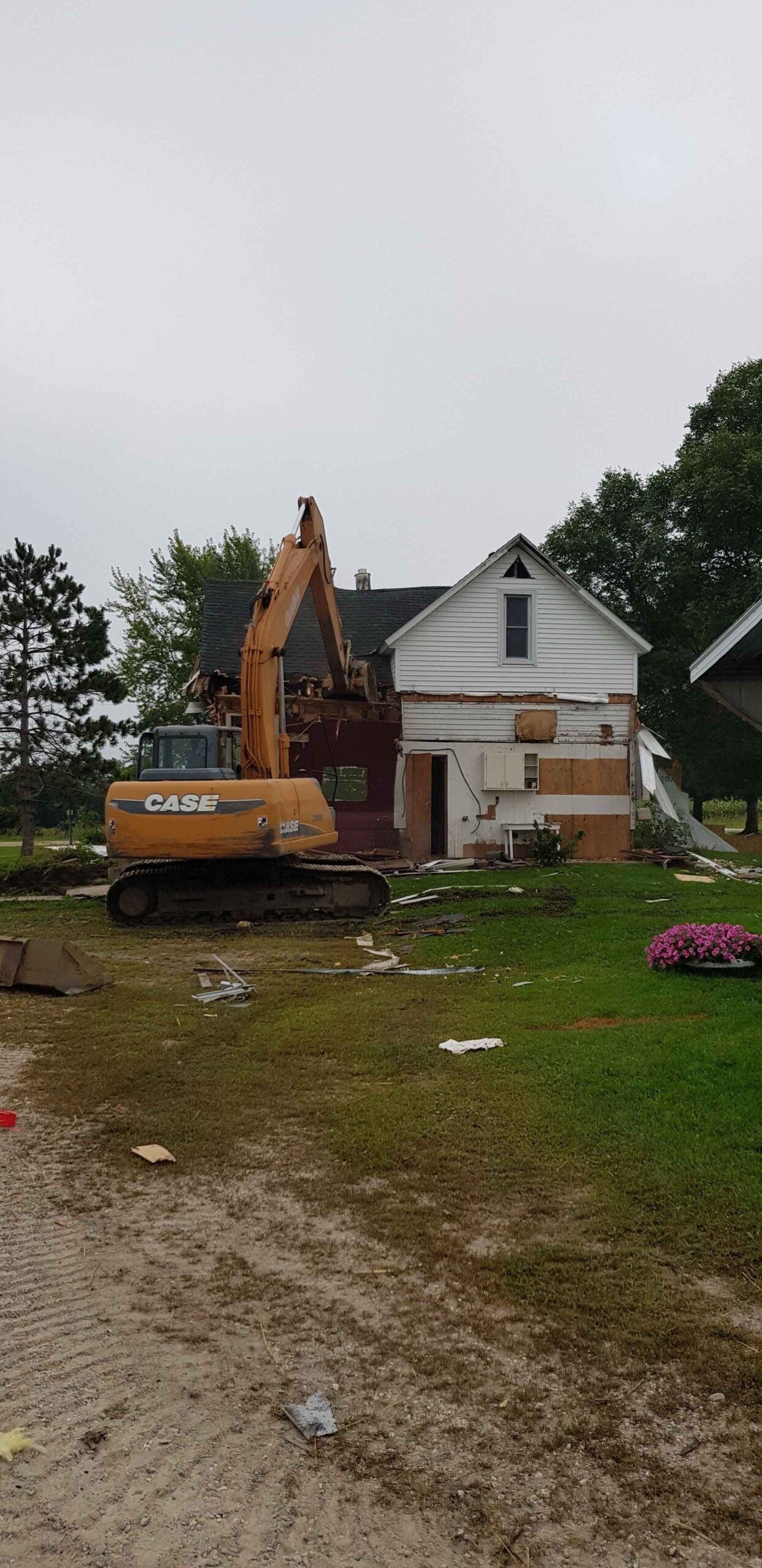 Excavator beside a white house on a muddy lot with scattered debris and trees under gray sky