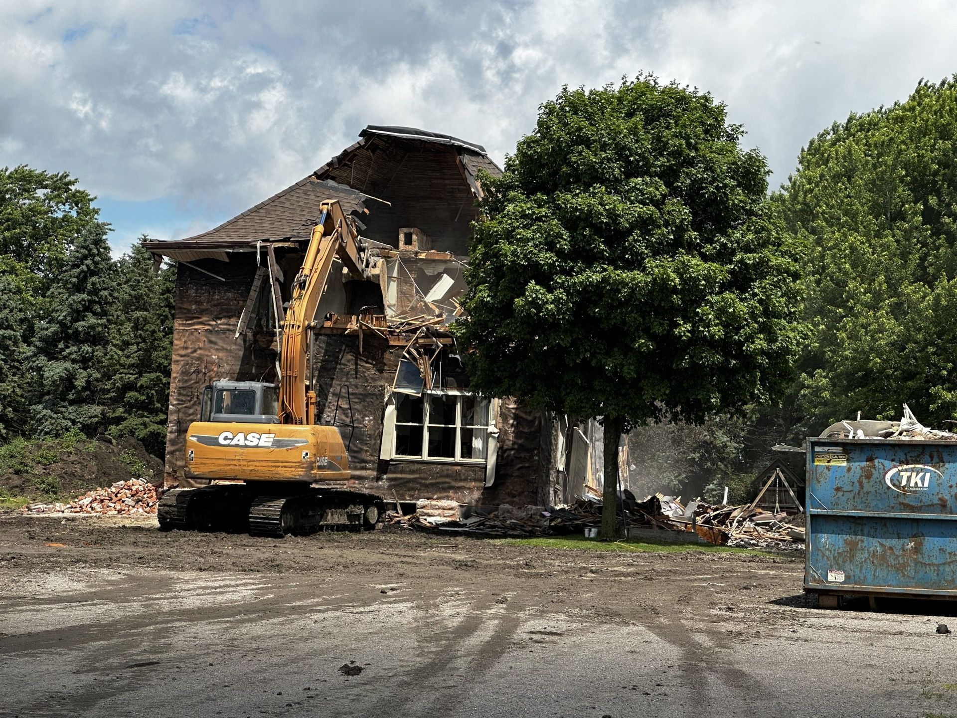Excavator demolishing a damaged house beside trees, with a blue dumpster in the foreground.