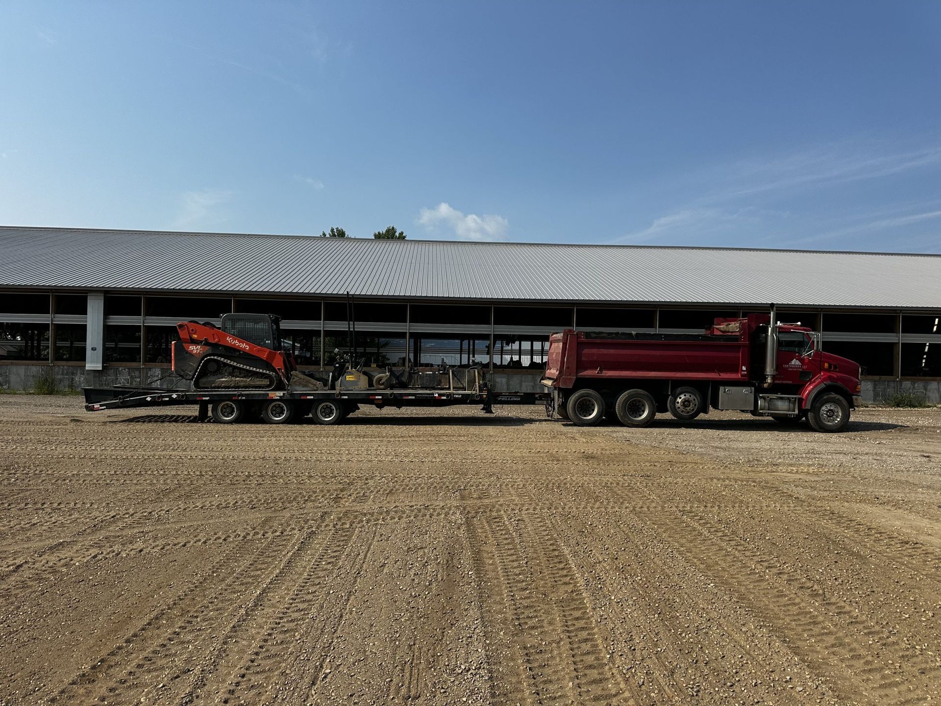 Red dump truck and orange excavator parked in front of a long building on a gravel lot under a blue sky