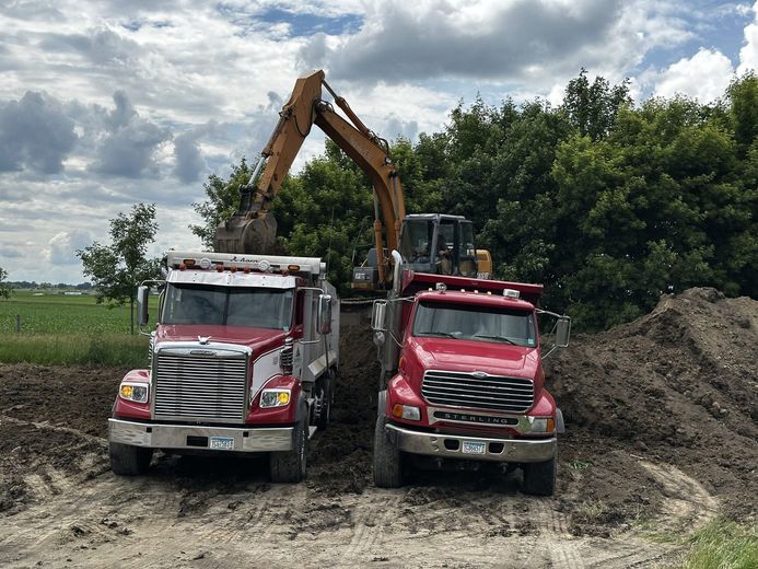 Two red dump trucks beneath an excavator at a muddy construction site under cloudy skies