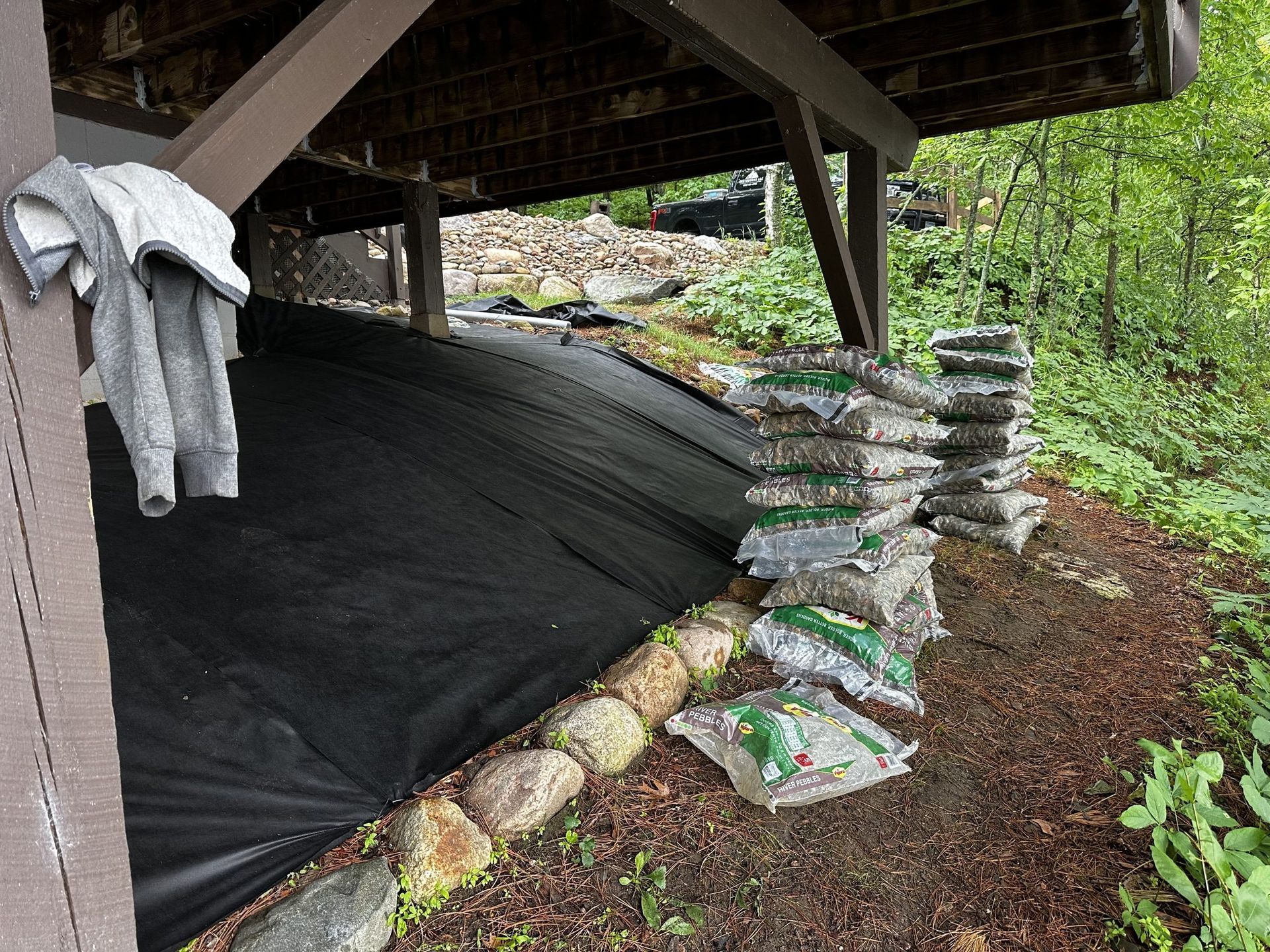 Black weed barrier under a raised structure, with stacked stone bags and a jacket hanging on the left