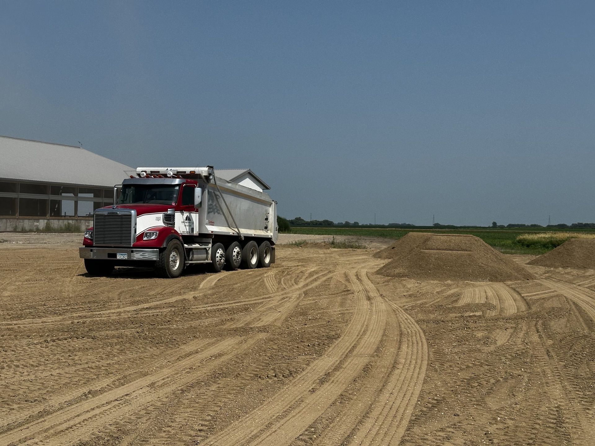 Red and white dump truck on a dirt construction site with tire tracks and low buildings in the background