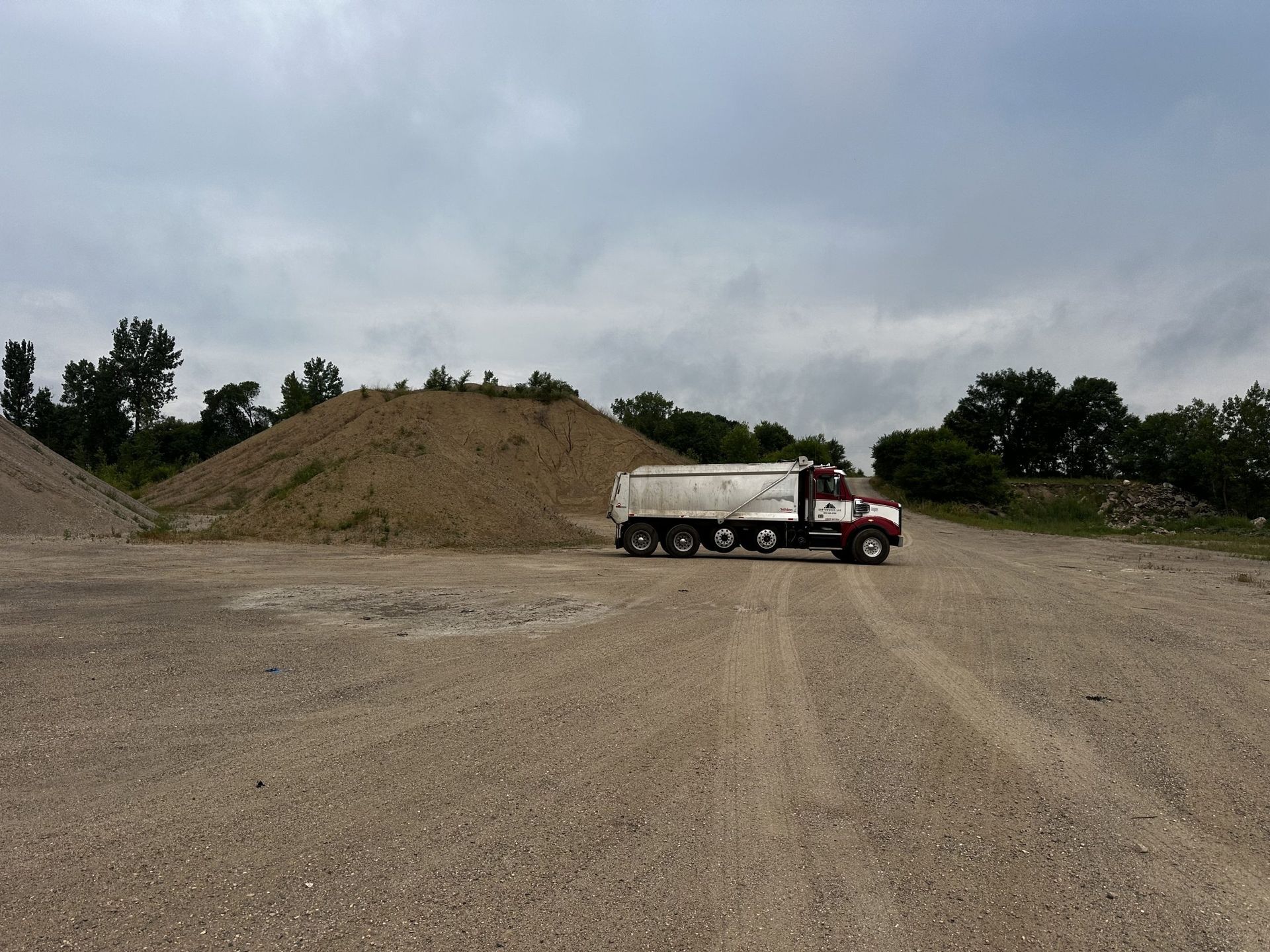 Dump truck parked on a gravel lot beside large dirt piles under a cloudy sky