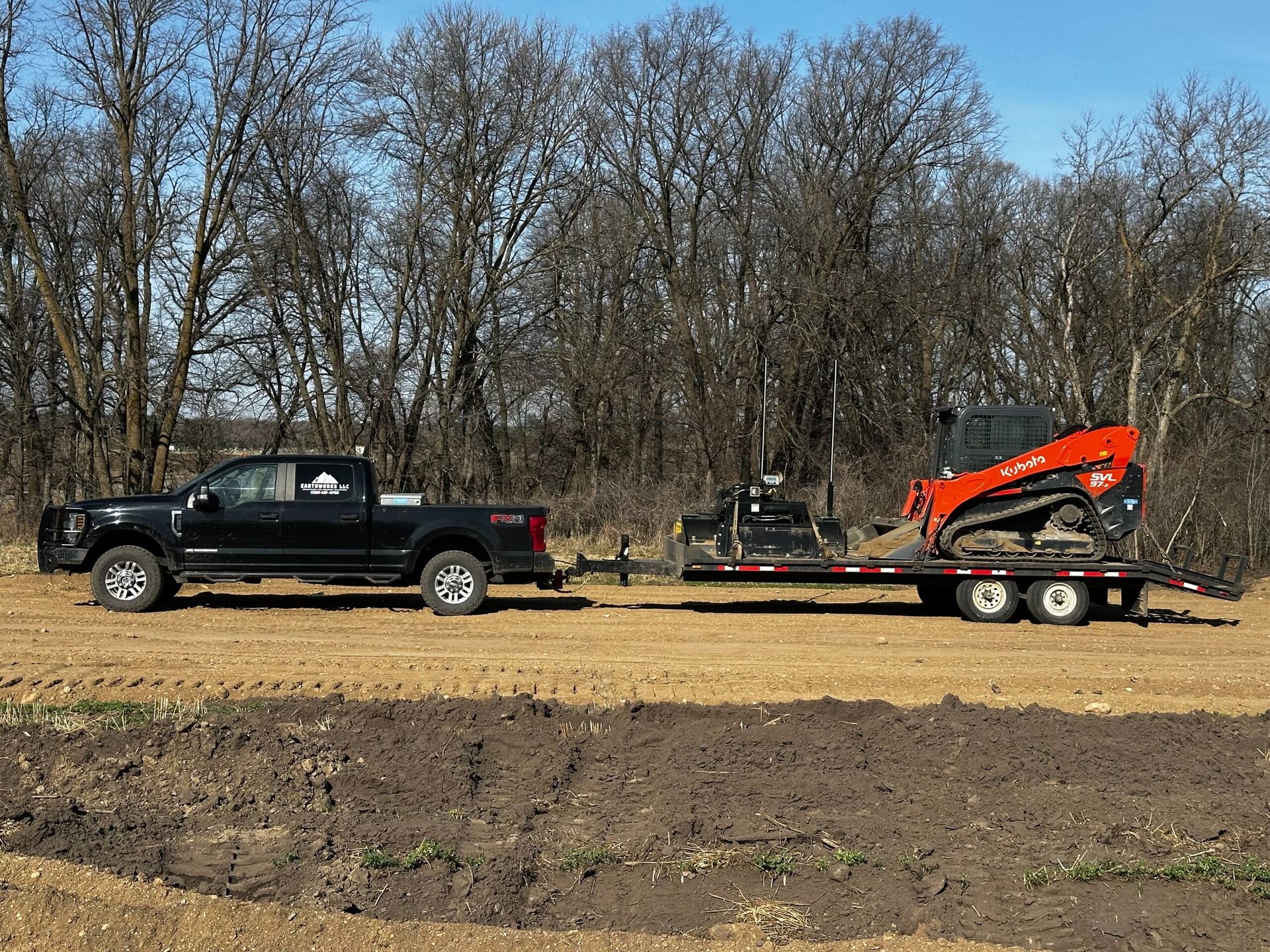Black pickup towing a trailer with an orange mini excavator across a dirt field under bare trees
