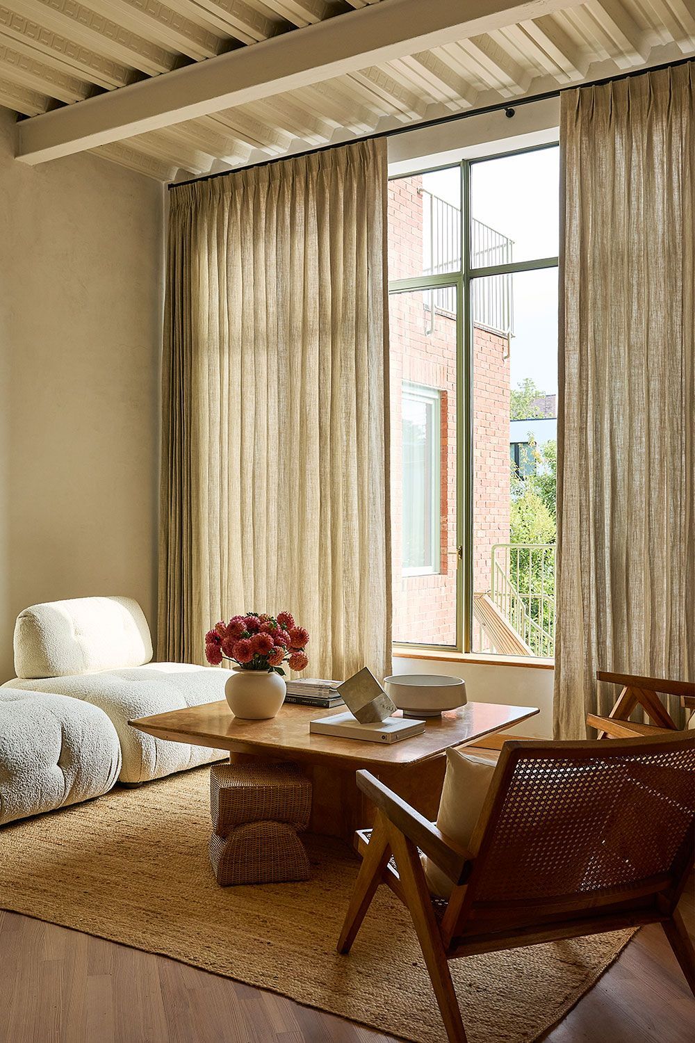 Modern living room with white sliding blinds, beige furniture, wood coffee table, and a view of trees outside.