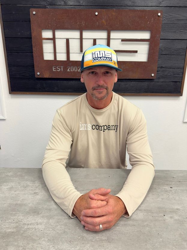 Man seated at a desk in front of an HHS sign, wearing a beige shirt and yellow cap, hands clasped.
