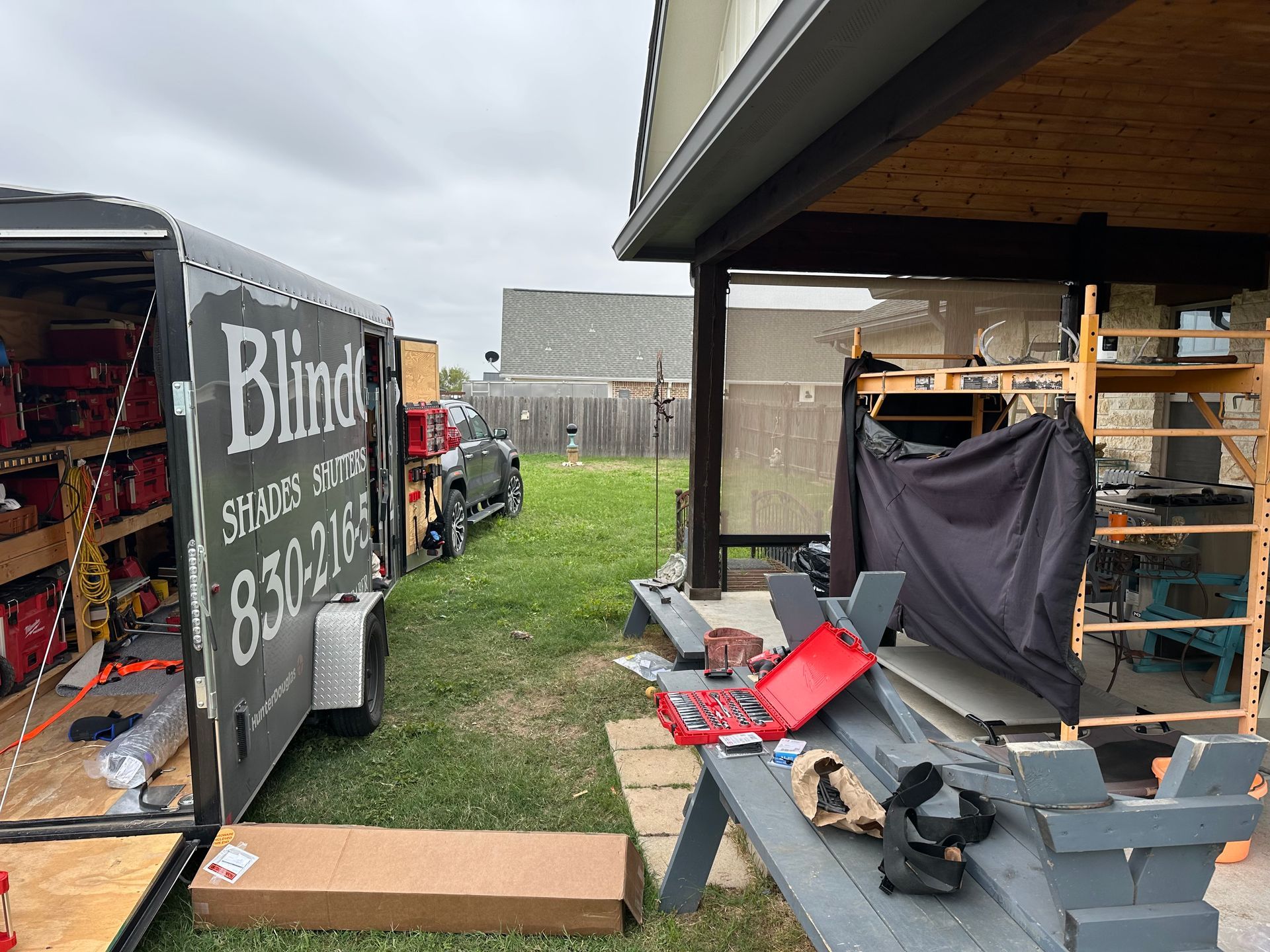 Outdoor patio and lawn with a black trailer labeled “Blind,” shelving, boxes, and construction supplies under an overhang