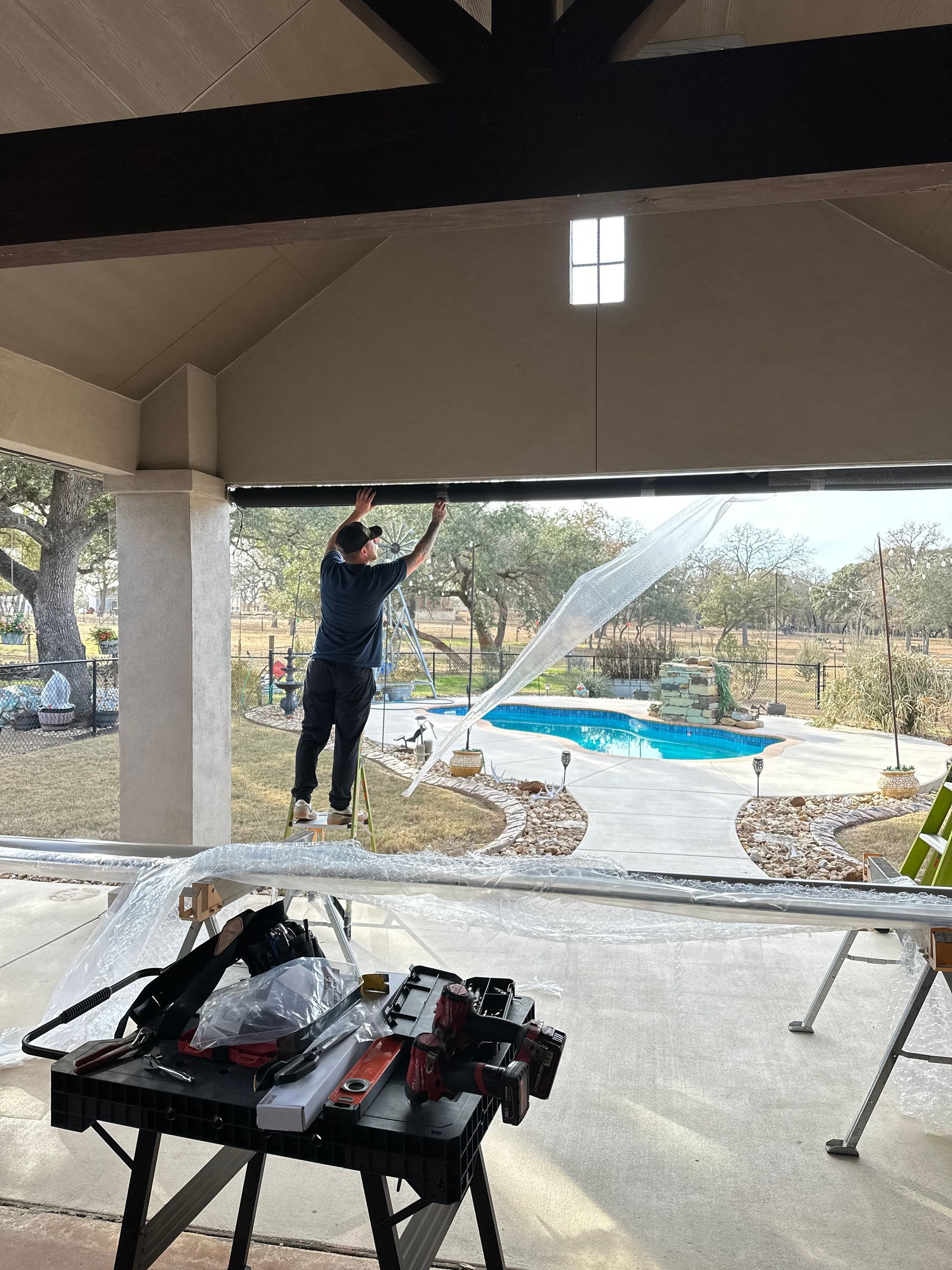 Person hanging a ceiling cable in an open-air patio with a ladder and tools nearby