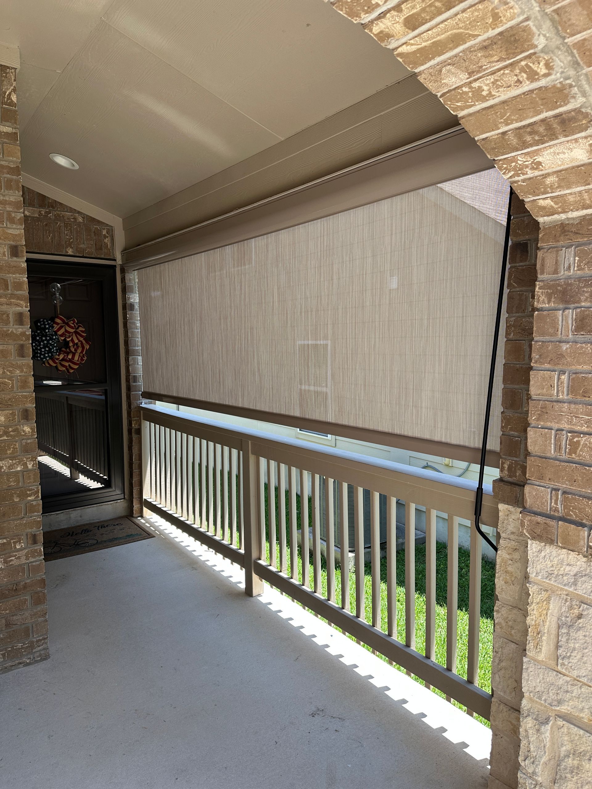 Covered apartment balcony with beige privacy shade, white railing, and brick posts overlooking greenery