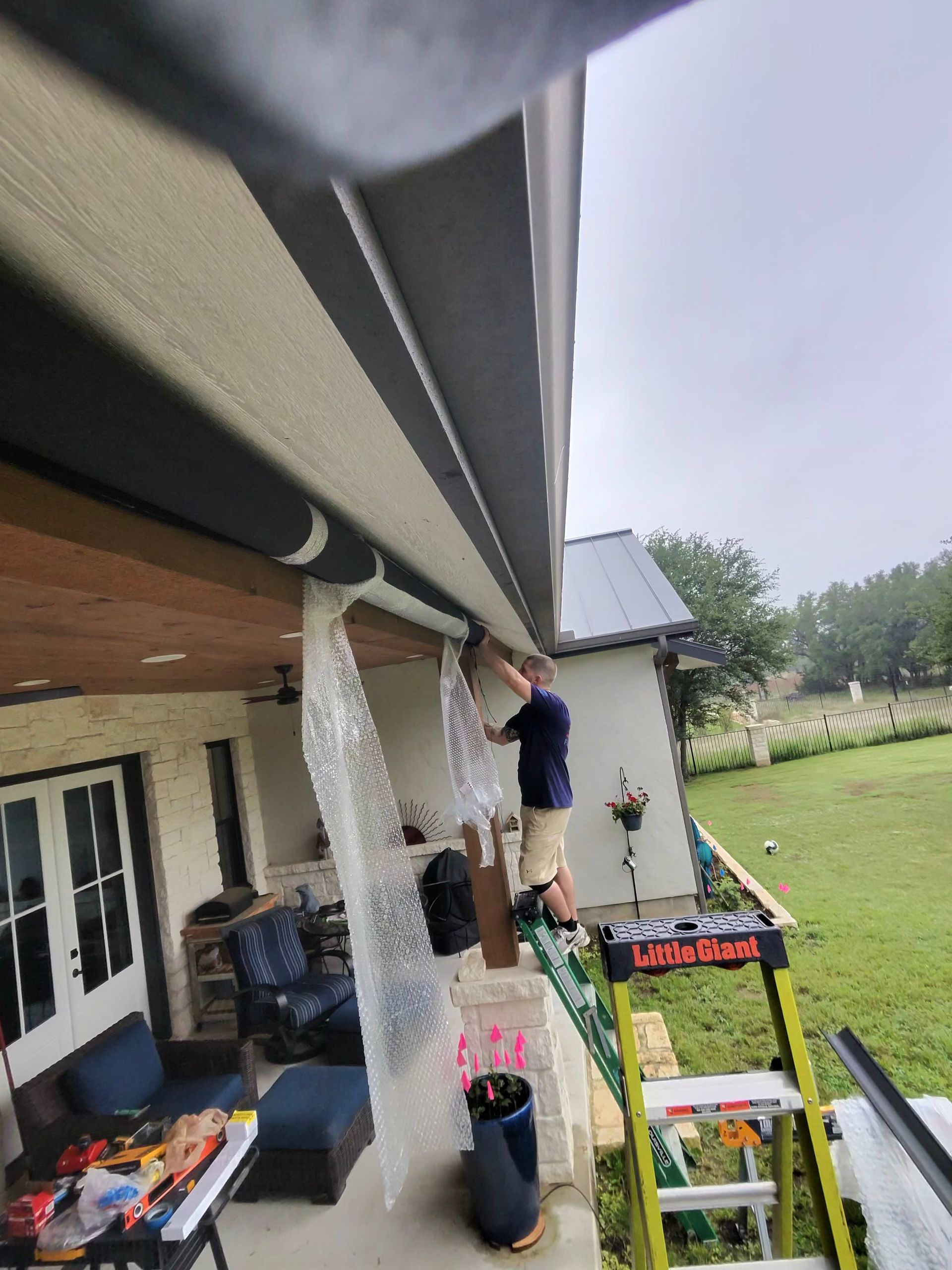 Person on a porch working near the roof gutter, with tools and materials beside a house and grassy yard.