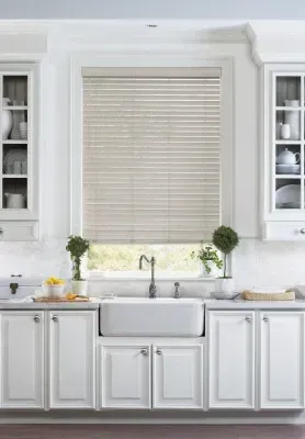 Bright white kitchen with a farmhouse sink, marble counters, and a window with closed blinds.